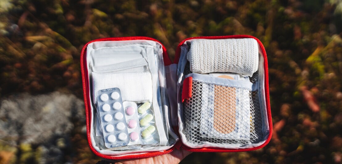 Hand hält eine geöffnete rote Erste-Hilfe-Tasche mit Verbandsmaterial, Pflastern und Medikamenten in Blisterpackungen.