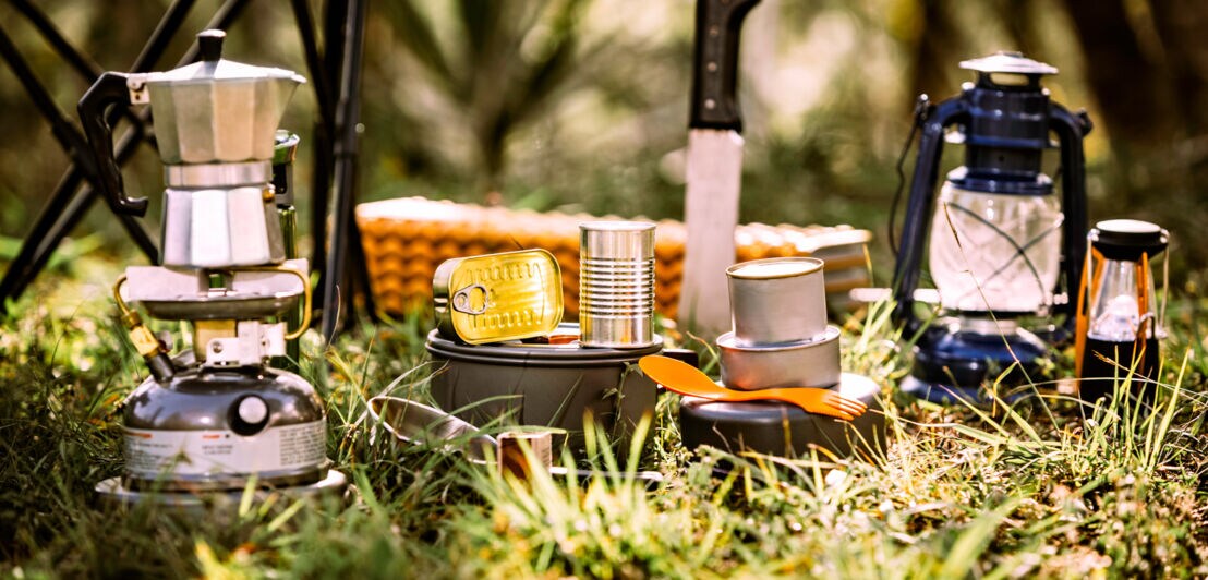 Camping-Kochutensilien auf Gras, darunter Gaskocher mit Espressokocher, Konservendosen, Messer, Pfanne, Laternen und Korb im Hintergrund.