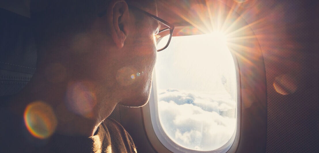 Mann mit Brille blickt aus dem Flugzeugfenster auf die Sonne und Wolken