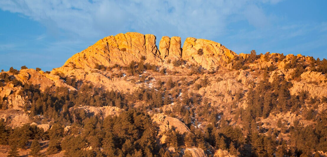 Felsiger Berg mit orangefarbenem Sonnenlicht und bewaldeten Hängen unter blauem Himmel mit Wolken