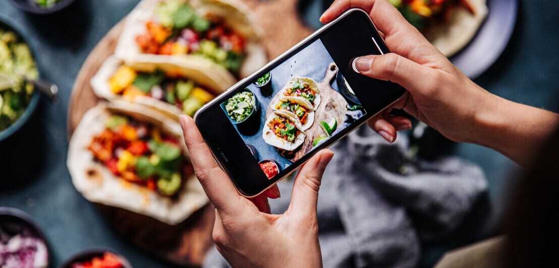 Person fotografiert mit Smartphone drei gefüllte Tacos auf einem Holzbrett, umgeben von kleinen Schalen mit Salsa und Guacamole