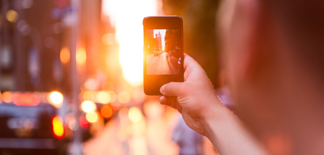 Person hält Smartphone und fotografiert eine belebte Straße bei Sonnenuntergang mit verschwommenem Hintergrund