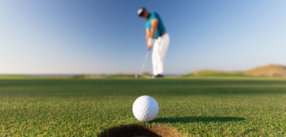 Nahaufnahme eines Golfballs an einem Loch auf einer Rasenfläche, im Hintergrund ein Mann mit Golfschläger in der Unschärfe vor blauem Himmel.
