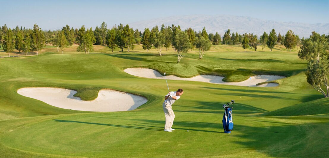 Ein Mann beim Abschlag auf einem hügeligen Golfplatz mit Sandbunkern bei Sonnenschein.