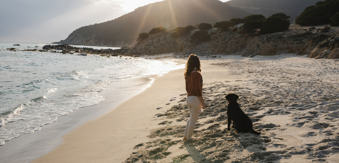 Frau und schwarzer Hund stehen am Strand und blicken auf das Meer bei Sonnenuntergang, Hügel im Hintergrund