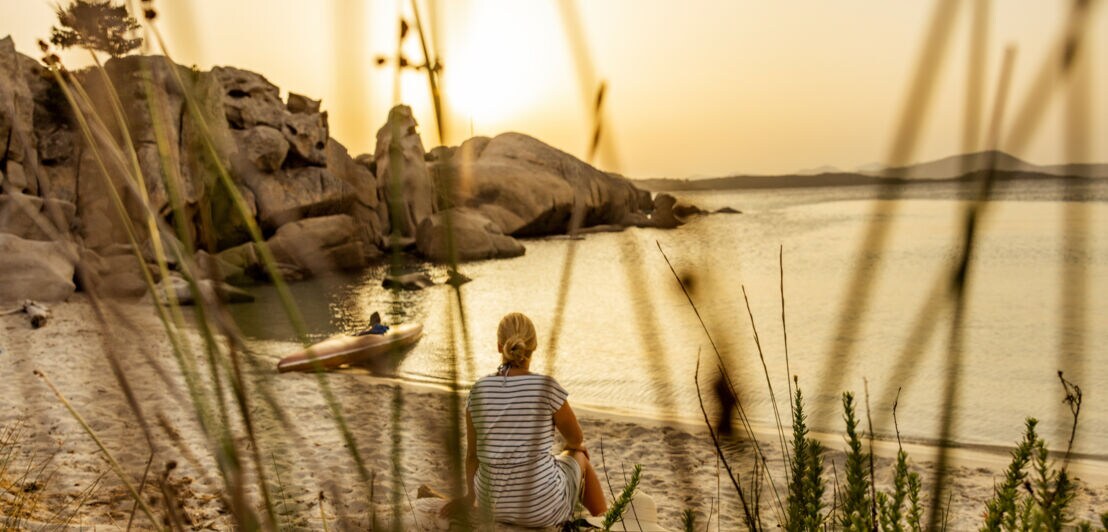 Person sitzt am Ufer eines Sees oder Meeres bei Sonnenuntergang, umgeben von Gras und Felsen