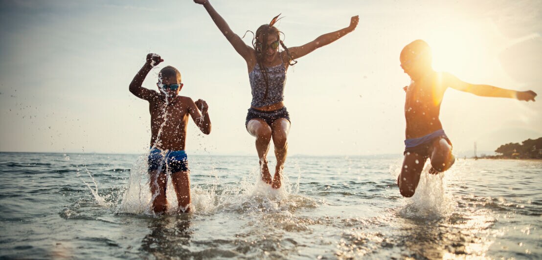 Drei Kinder springen lachend im flachen Wasser am Strand bei Sonnenuntergang