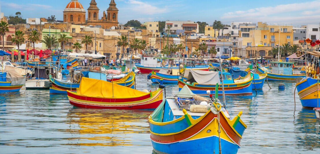 Bunte traditionelle Fischerboote im Hafen von Marsaxlokk, Malta, mit Kirche und Gebäuden im Hintergrund.