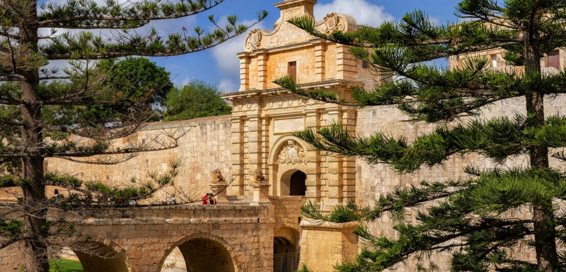 Historisches Tor mit Brücke und Steinbögen, umgeben von grünen Bäumen unter blauem Himmel.