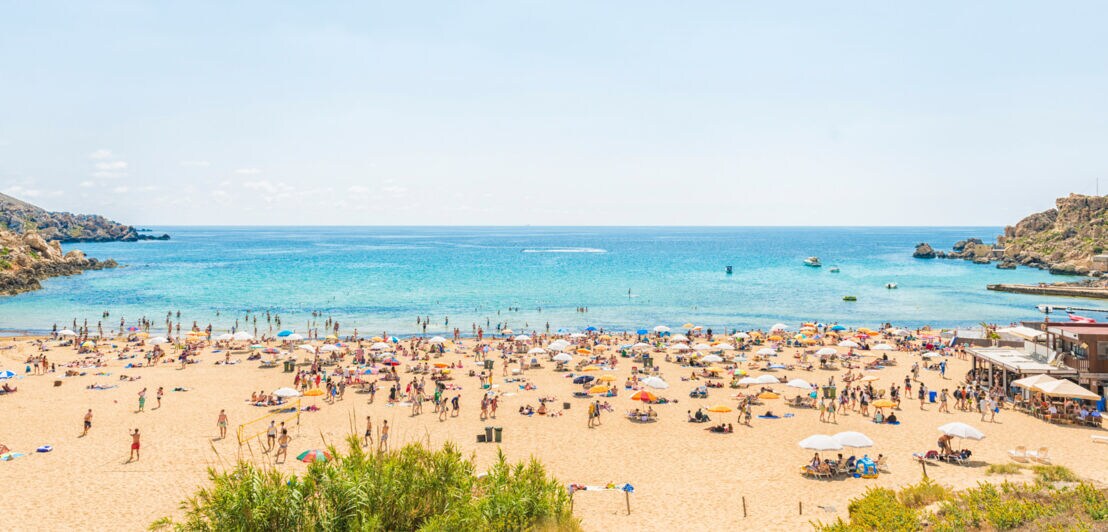 Strand mit vielen Menschen, Sonnenschirmen und klarem blauem Meer unter hellem Himmel