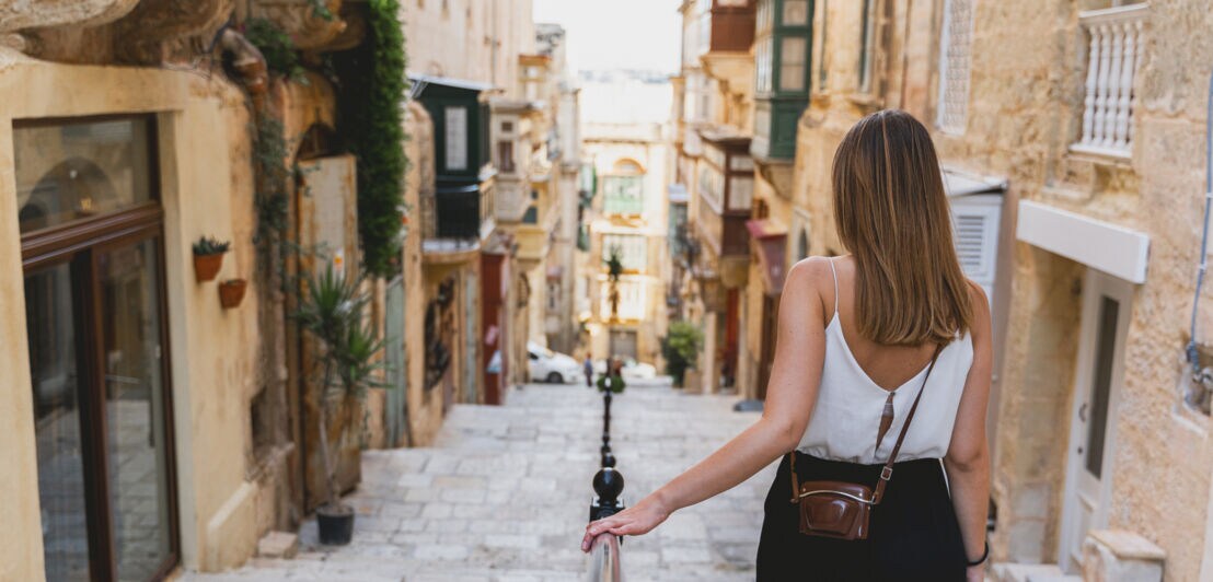 Frau mit weißem Top und schwarzer Hose geht eine steinerne Treppe in einer engen Gasse mit historischen Gebäuden hinunter