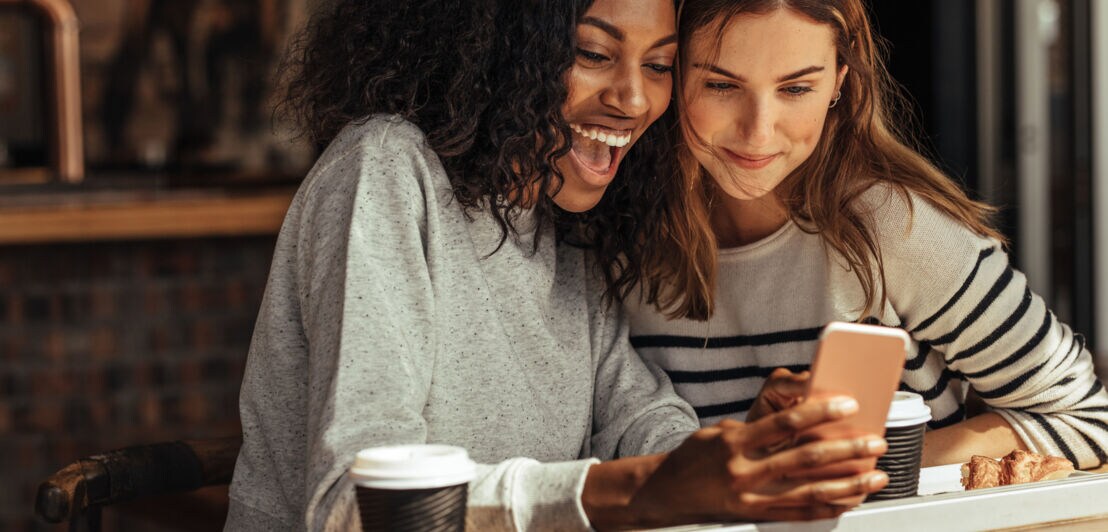 Zwei junge lächelnde Frauen schauen gemeinsam auf ein Smartphone mit Kaffeebechern vor sich.