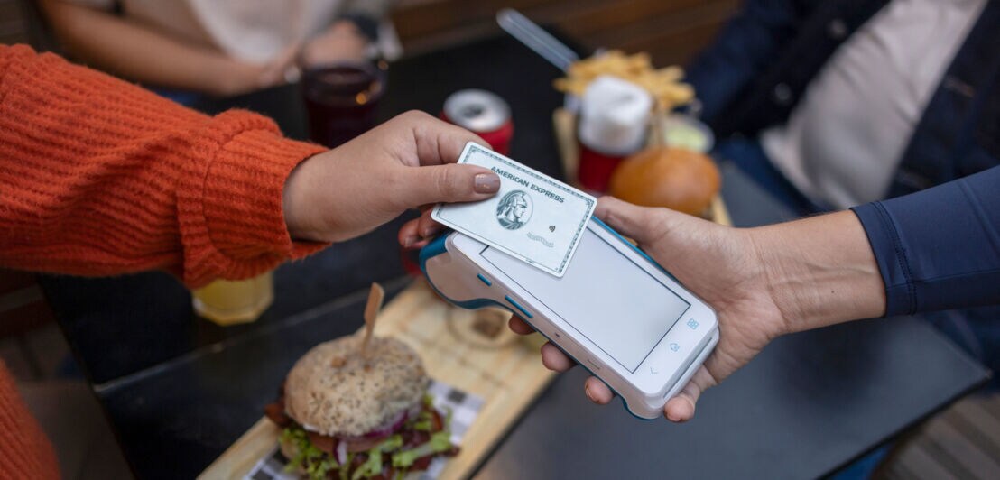 Eine Person hält eine silberne Kreditkarte von American Express an ein Kartenlesegerät, das eine andere Person über einen Tisch mit Hamburgern hält.