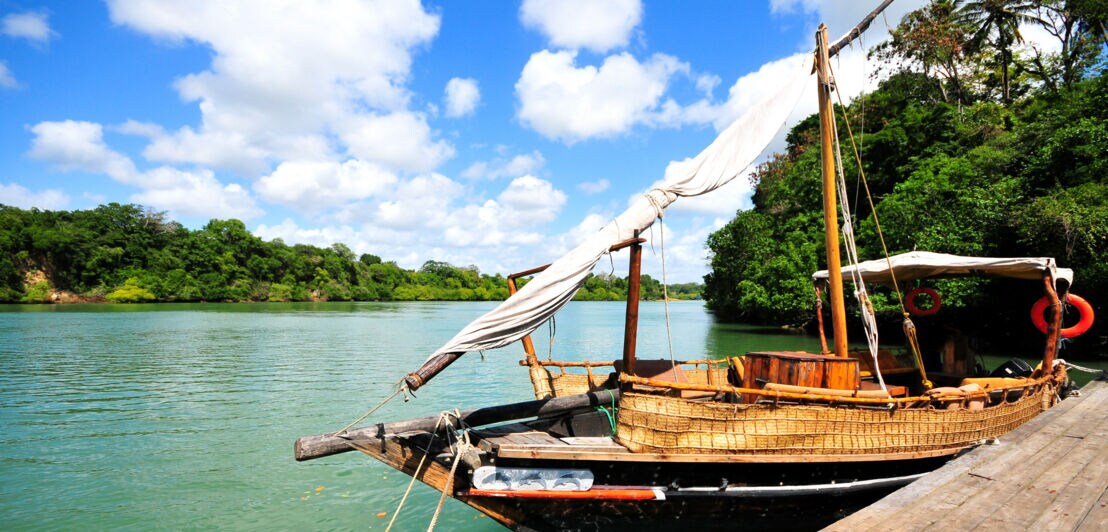 Traditionelles Holzsegelboot mit gefalteten weißen Segeln am Ufer eines ruhigen Flusses, umgeben von grünen Bäumen unter blauem Himmel mit Wolken