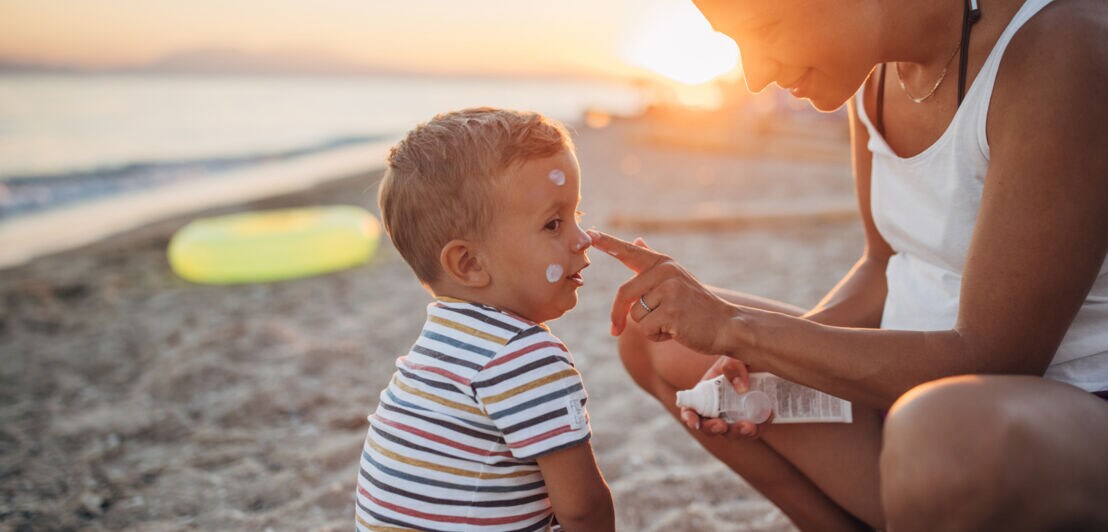 Frau trägt Sonnencreme auf die Nase eines kleinen Jungen am Strand bei Sonnenuntergang auf