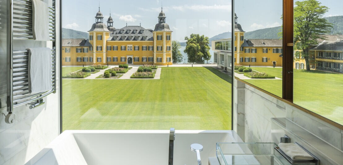 Aussicht auf eine Rasenfläche vor einem gelben Schlosshotel an einem See durch ein Fenster in einem modernen Badezimmer.
