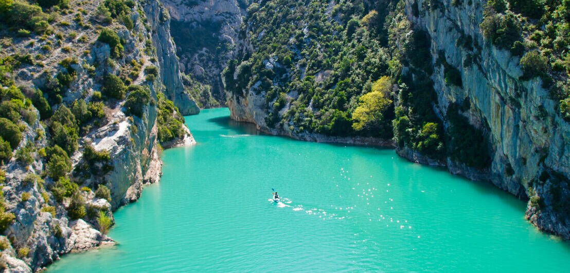 Türkisfarbener Fluss fließt durch enge, bewaldete Felsenschluchten unter klarem Himmel, eine Person paddelt im Kajak