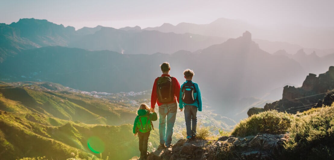 Drei Personen mit Rucksäcken wandern auf einem Bergpfad mit Blick auf ein Tal und entfernte Bergketten im Sonnenlicht