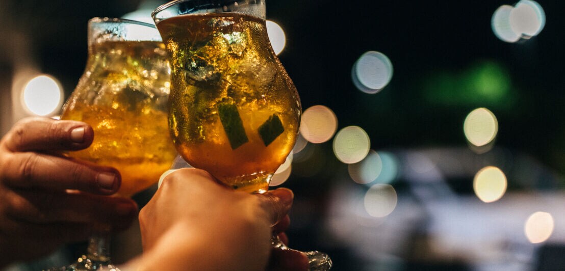 Zwei Hände stoßen mit Gläsern voller Eis und goldfarbenem Getränk bei Nacht an