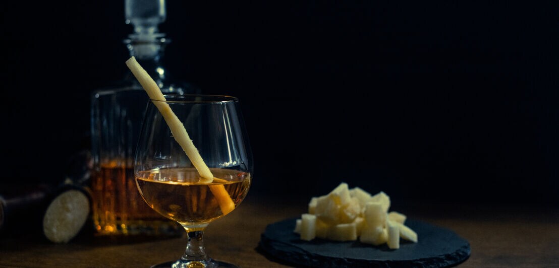 Glas mit braunem Getränk und Zuckerrohr, daneben ein Teller mit Zuckerrohr und eine Karaffe im Hintergrund