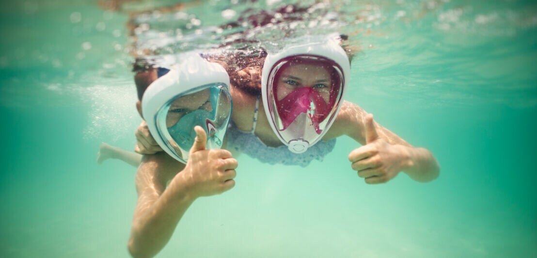 Zwei Kinder mit Vollgesichtsschnorchelmasken tauchen unter Wasser und zeigen Daumen hoch