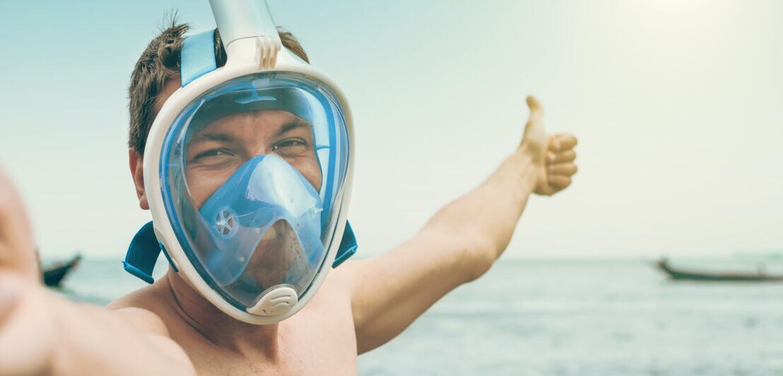 Mann mit Vollgesichts-Schnorchelmaske am Meer, der Daumen hoch zeigt