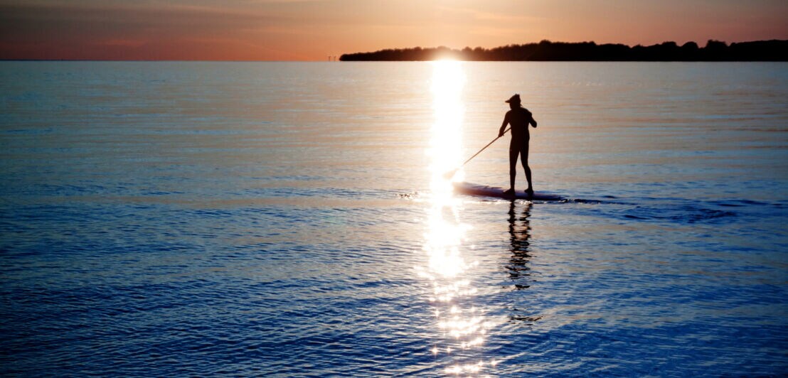 Person beim Stand-up-Paddling auf ruhigem Wasser bei Sonnenuntergang mit Sonnenlichtspiegelung