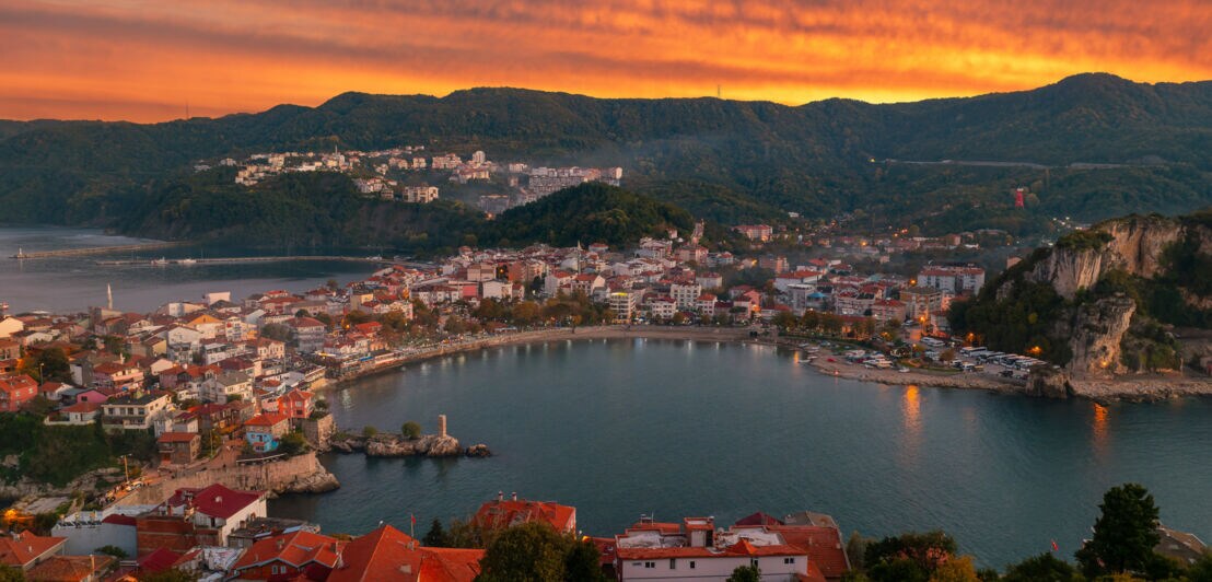 Bucht mit Hafen und roten Dächern vor bewaldeten Hügeln bei Sonnenuntergang mit orangefarbenem Himmel.