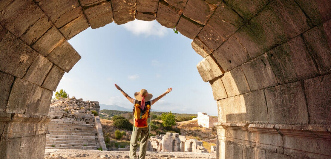 Person mit Hut und Rucksack steht mit ausgebreiteten Armen vor antiken Ruinen, gesehen durch einen steinernen Bogen.