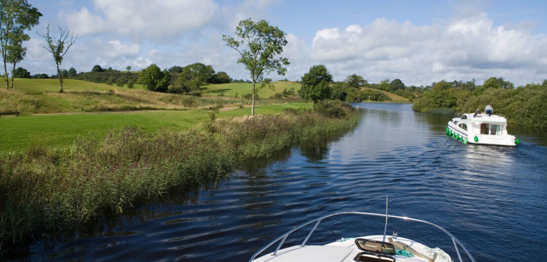 Zwei moderne Hausboote fahren auf einem ruhigen Fluss durch eine grüne Wiesenlandschaft mit Schilfrohr.