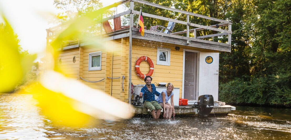 Ein fröhliches Paar sitzt am Rande eines fahrenden Hausbootes auf einem Fluss und spritzt mit Wasser.