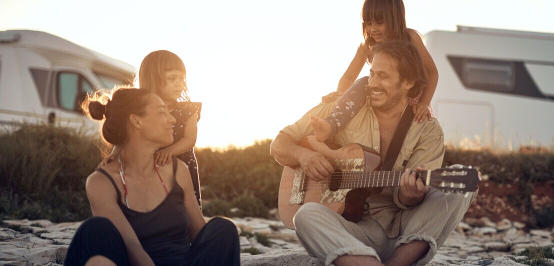 Familie mit zwei Kindern sitzt auf Felsen bei Sonnenuntergang, Vater spielt Gitarre, Wohnmobile im Hintergrund