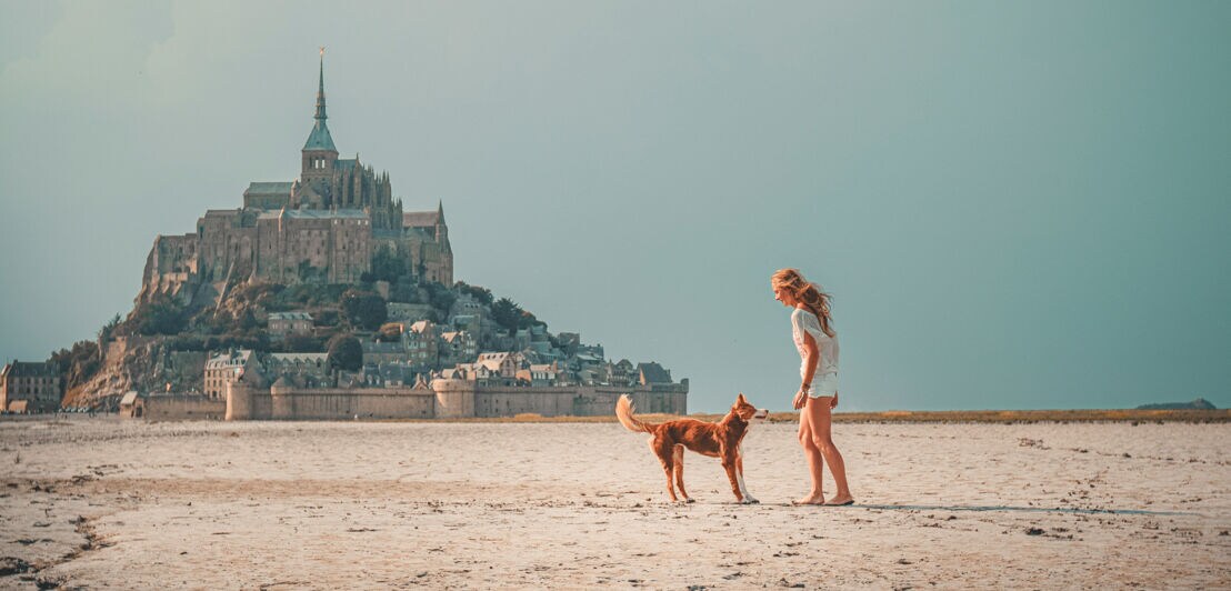 Frau in weißem Outfit spielt mit braunem Hund auf sandigem Boden vor Mont-Saint-Michel im Hintergrund.