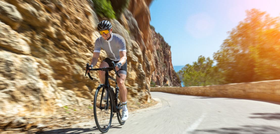 Mann in Radfahrkleidung und Helm fährt Rennrad auf kurviger Bergstraße mit Felswand und Bäumen im Hintergrund bei Sonnenschein