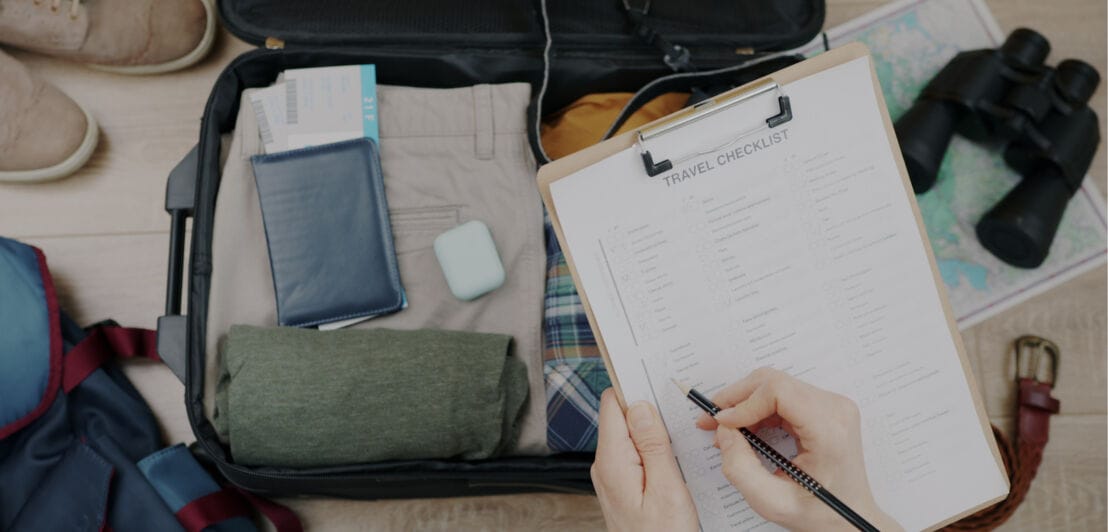 Offener Koffer mit Kleidung, Reisepass, AirPods und Fernglas, daneben Rucksack und Schuhe, Person füllt Packliste aus