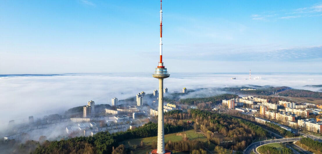 Hoher Fernsehturm mit rot-weiß gestreifter Spitze vor einem nebelverhangenen Stadtpanorama.