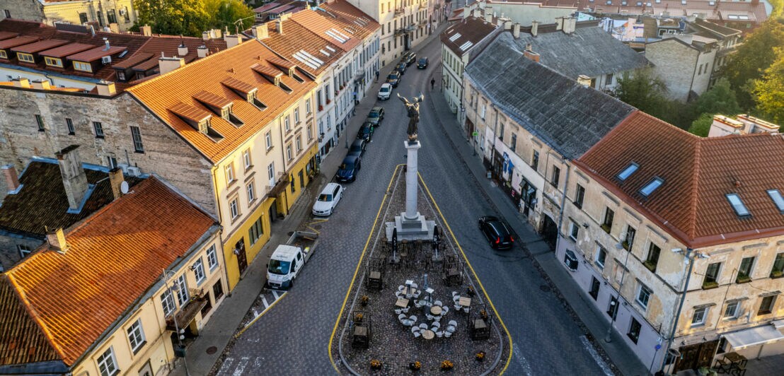 Luftaufnahme eines Straßenzuges mit einer Verkehrsinsel mit Siegessäule und Sitzplätzen in einem Altstadtviertel.