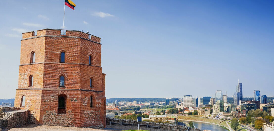 Eckiger Burgturm aus rotem Backstein mit litauischer Flagge auf einer Festung auf einem Hügel vor Stadtpanorama.
