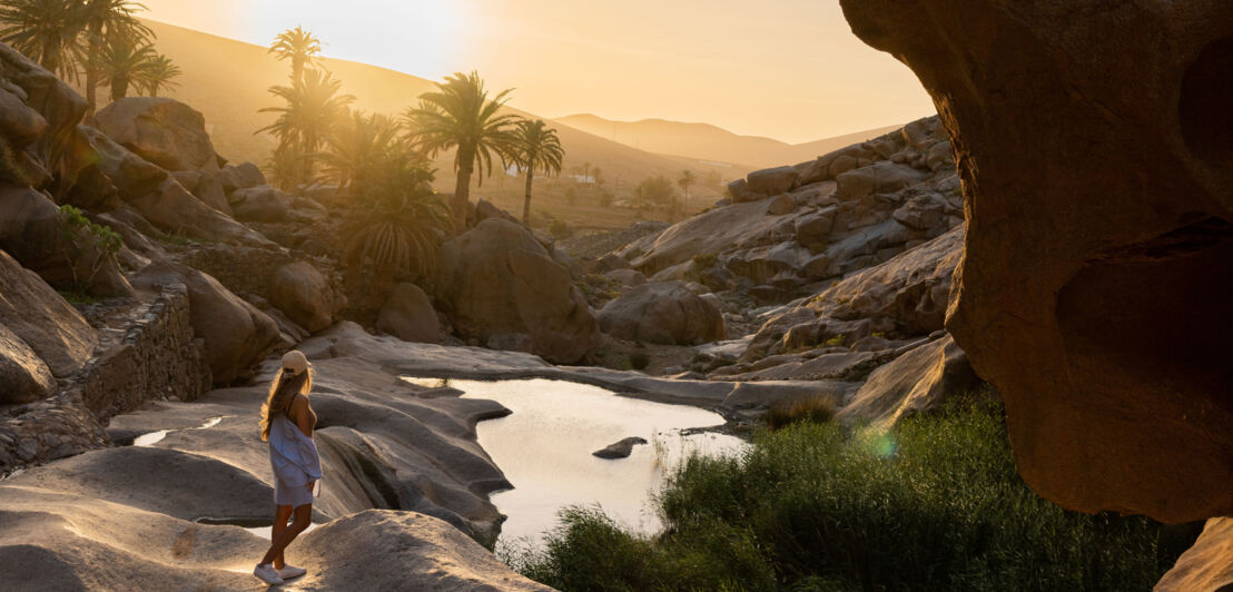 Eine Frau steht in einer Felslandschaft aus vulkanischem Gestein mit Palmen bei Sonnenuntergang.