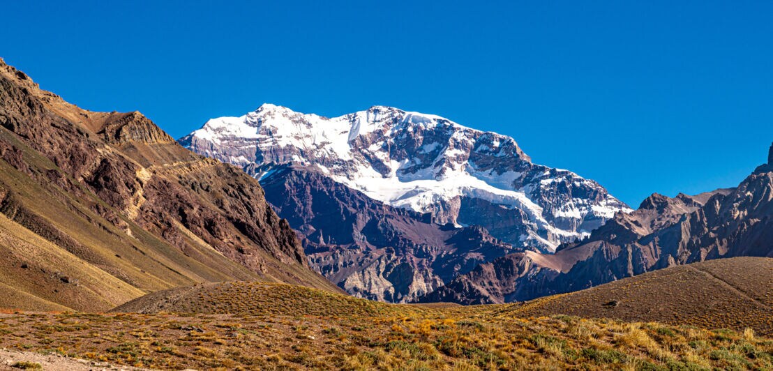 Schneebedeckter Berggipfel vor klarem blauem Himmel, umgeben von felsigen Hügeln und trockenem Grasland