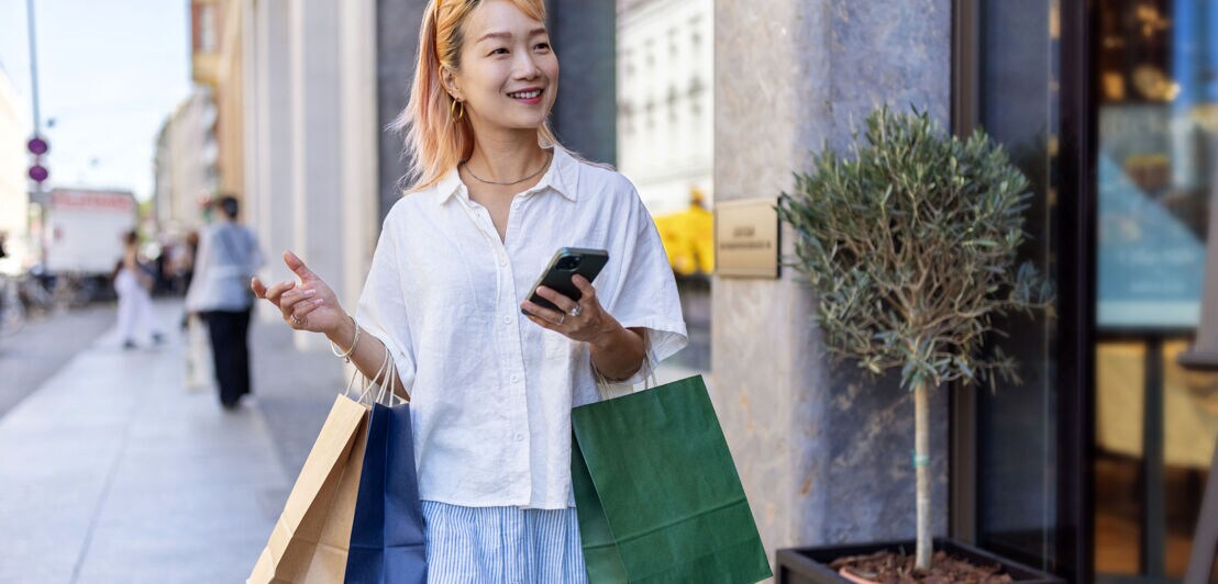 Eine junge asiatische Frau mit blonden Haaren mit Einkaufstüten und Smartphone in den Händen geht lächelnd über eine Einkaufsstraße.