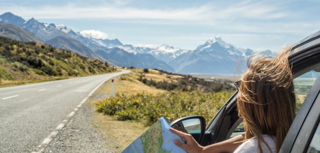 Frau schaut aus Autofenster auf eine Landkarte mit Blick auf eine offene Straße und schneebedeckte Berge im Hintergrund.