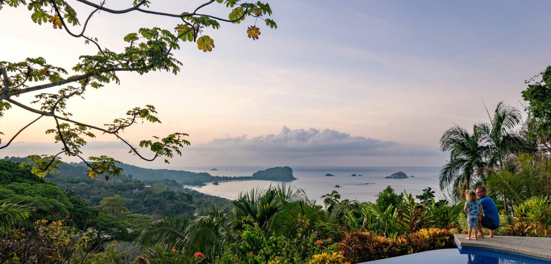 Blick auf tropische Küstenlandschaft mit Inseln im Meer, Pflanzen und Infinity-Pool im Vordergrund, zwei Personen am Poolrand.