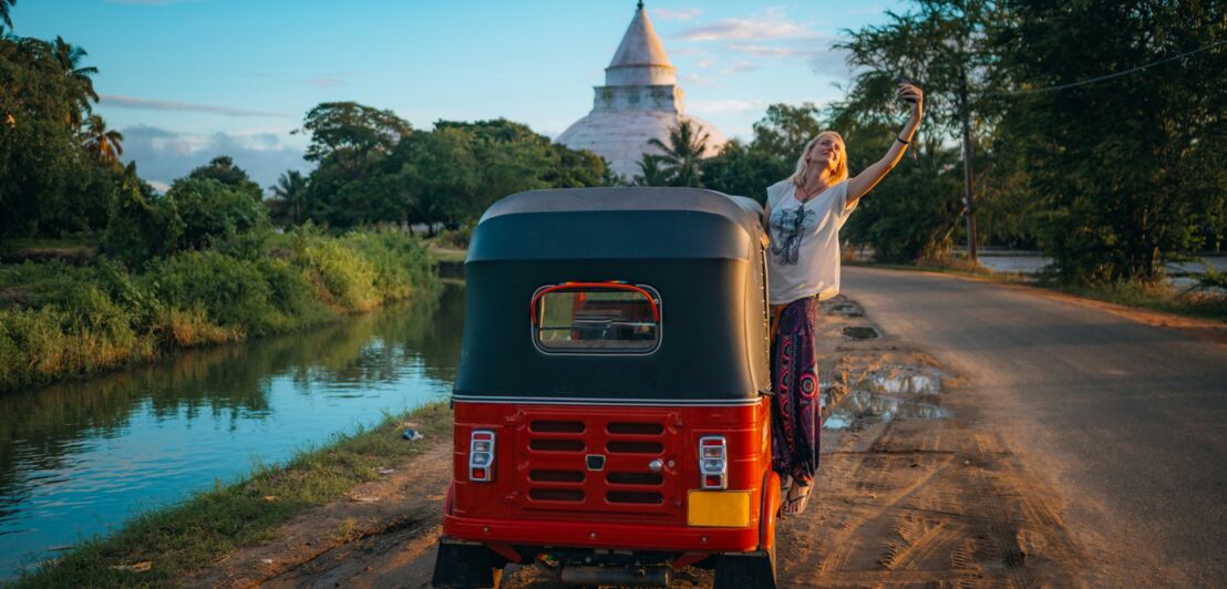Frau macht Selfie neben rotem Tuk-Tuk auf ländlichem Weg mit Tempel im Hintergrund.