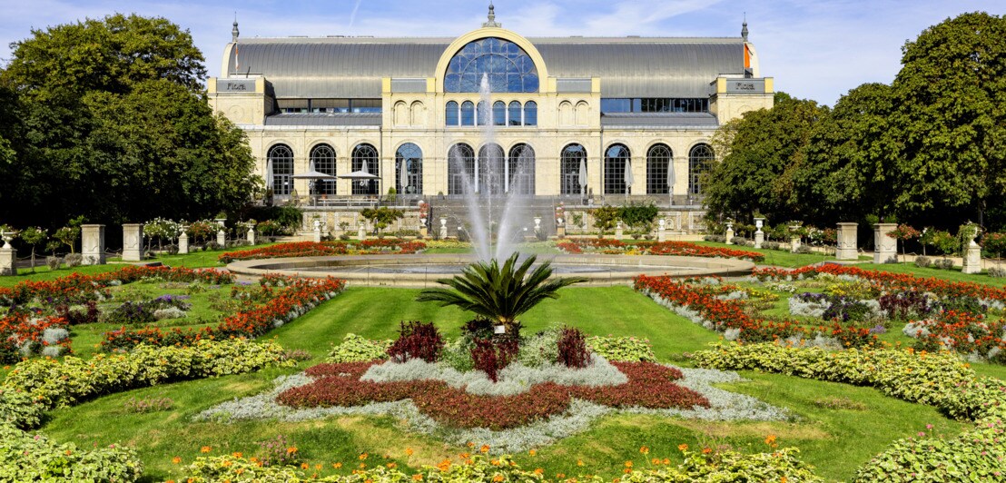 Symmetrisch angelegter botanischer Garten mit bunten Blumenbeeten und Springbrunnen vor einem historischen Gebäude mit großen Fenstern und Rundbogenfenster