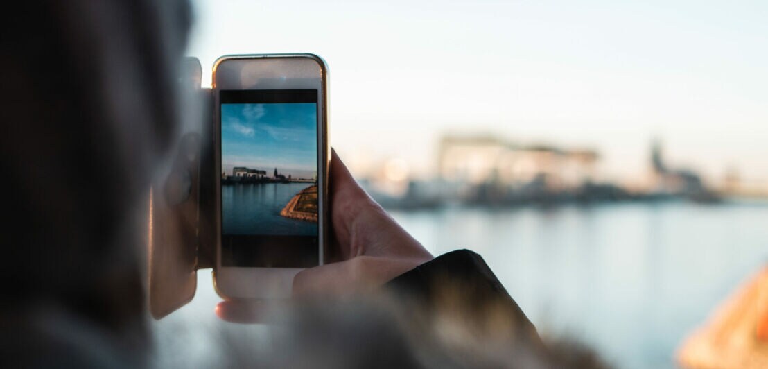 Person hält Smartphone und fotografiert eine Wasserlandschaft mit Hafen und Gebäuden im Hintergrund