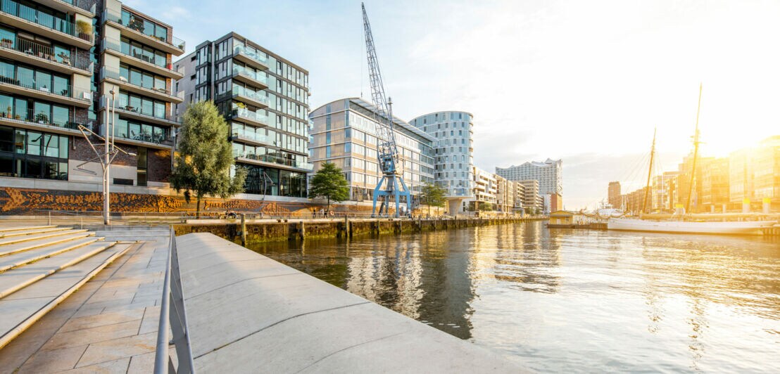 Moderne Wohngebäude am Ufer eines ruhigen Flusses bei Sonnenuntergang mit einer blauen Hafenkran-Skulptur.