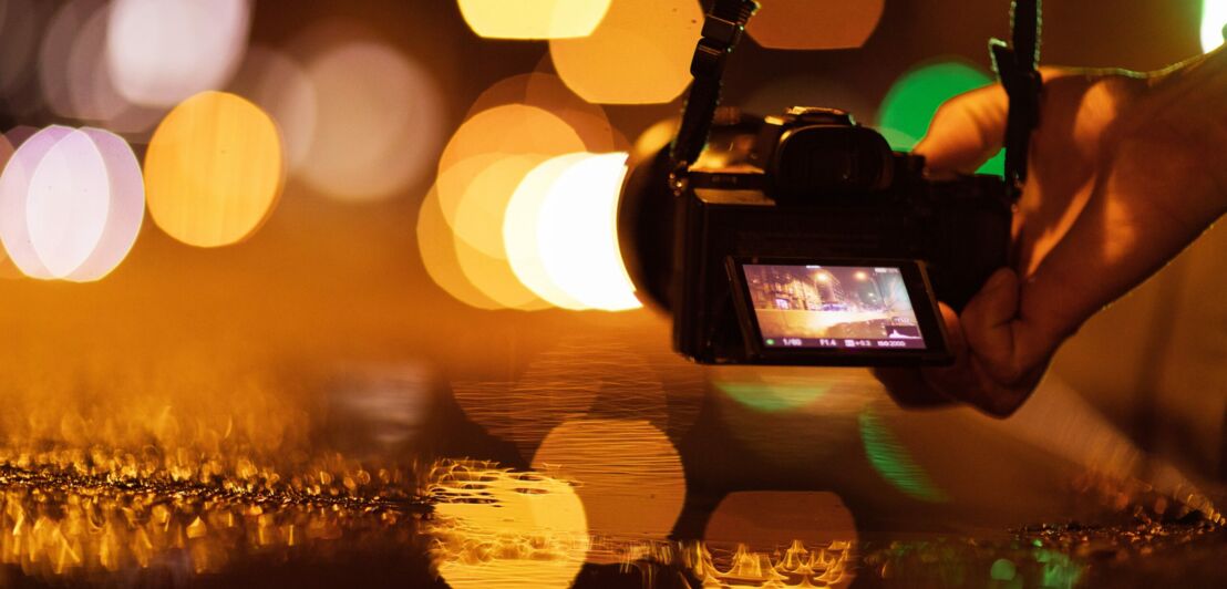 Digitalkamera mit Display, das eine nächtliche Straßenszene mit Lichtern zeigt, vor unscharfem Hintergrund mit orangefarbenen Lichtpunkten