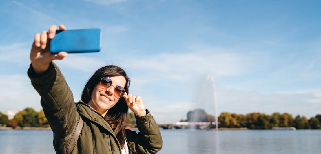 Eine Frau macht ein Selfie mit der Fontäne der Binnenalster im Hintergrund.
