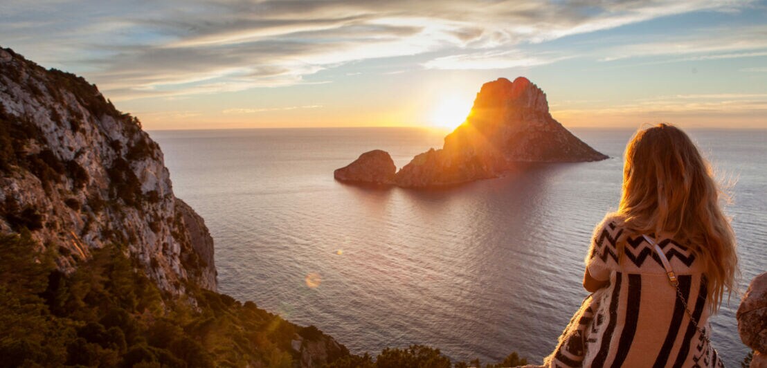 Frau mit langem Haar und gestreiftem Pullover sitzt auf Felsen und blickt auf Sonnenuntergang über Meer und Insel